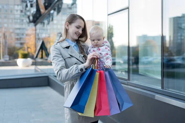 Šťastná mladá matka s malou dcerou na ramenou a nákupními taškami v ruce. Nákupní den. Nákupní centrum na pozadí — Stock fotografie