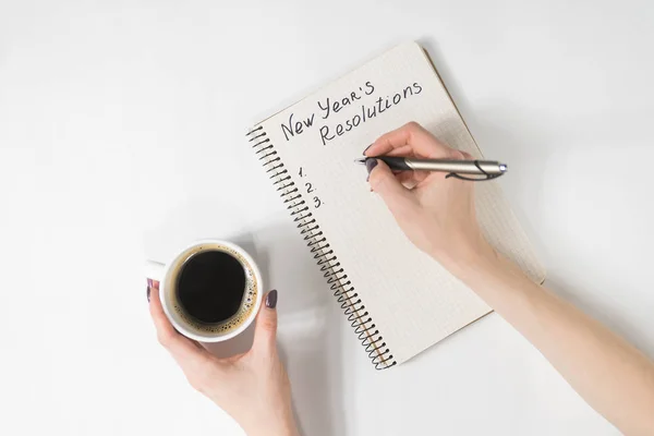 Phrase New Year 's resolutions in the notebook. Женская рука с ручкой и чашкой кофе — стоковое фото