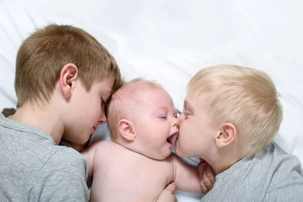 Dos hermanos mayores besan tiernamente y abrazan al niño más pequeño en una cama blanca. Familia feliz — Foto de Stock