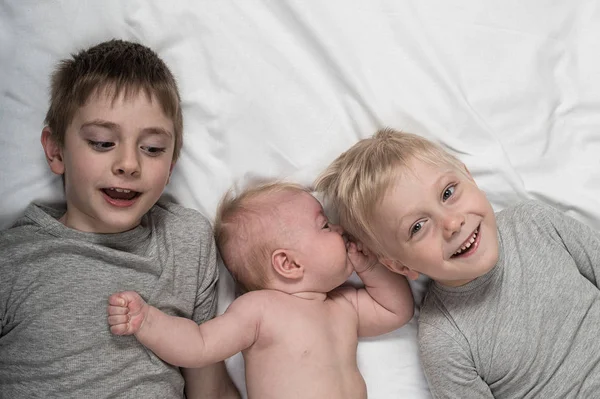 Two older brothers play and laugh with the youngest baby in a white bed. Happy childhood, big family — Stock Photo, Image