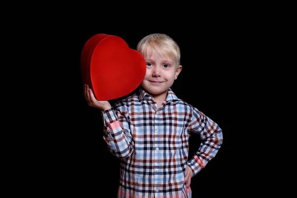 Menino se inclina contra sua bochecha caixa de forma de coração vermelho em um fundo preto — Fotografia de Stock