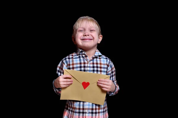 Lindo chico rubio sostiene un sobre con un corazón rojo. Felicidades, San Valentín. Fondo negro — Foto de Stock