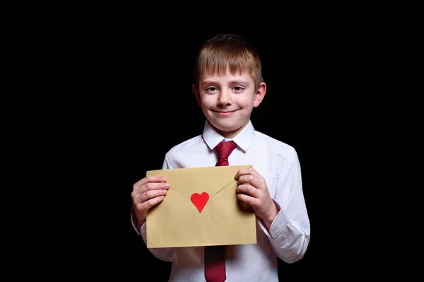Lindo chico con camisa y corbata sostiene un sobre con un corazón. Aislar sobre fondo negro — Foto de Stock
