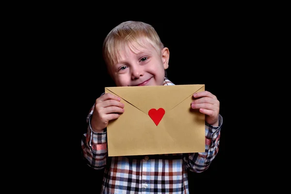Leuke glimlachende blonde jongen met een envelop met een rood hart. Gefeliciteerd, Valentijnsdag. Zwarte achtergrond — Stockfoto