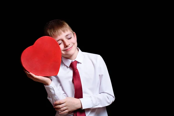 Closed eyes boy leans against his cheek red heart shape box on a dark background — 스톡 사진