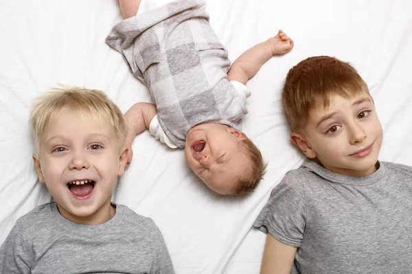 Dois irmãos mais velhos e o bebé mais novo numa cama branca. Infância feliz, família grande. Vista superior — Fotografia de Stock