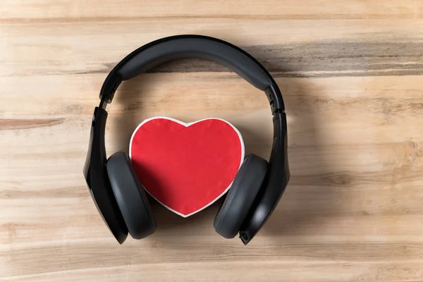 Full-size wireless headphones pulled over small red heart-shaped box on a light brown wooden table. Love music concept. Directly above — Stock Photo, Image