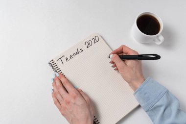 Eğilimler 2020 yazıtları. Notepad 'de yazan kadın elleri. Not defteri ve kahve fincanı beyaz arkaplanda..
