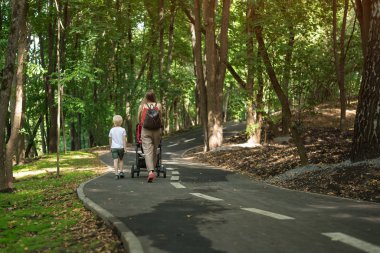 Çocuklu ve pusetli bir anne parkta yürüyor. Açık havada yürüyor. Arka plan