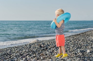 Küçük sarı saçlı çocuk deniz kıyısında elinde şişme yüzükle duruyor. Çocuk denize bakıyor.