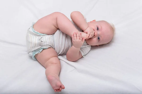 Pequeño bebé mordisquea su pie en la cama blanca. Lindo bebé de 3 meses. Gimnasia para el recién nacido —  Fotos de Stock