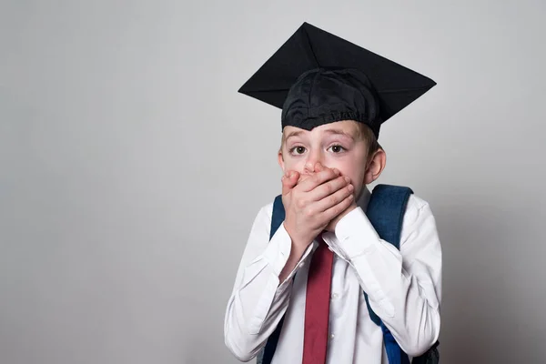 Student scared covered his mouth with his hands. Boy in school uniform and academic hat. White background.
