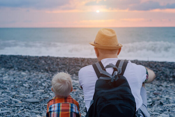 Молодой отец и маленький сын сидят на пляже и смотрят на море и закат. Вид сзади
.