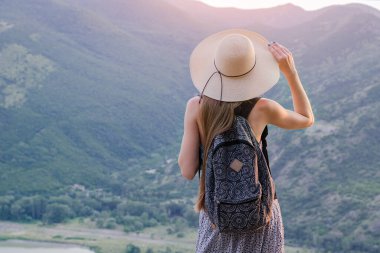 Genç bir kadın dağ vadisine bakıyor ve el şapkasını tutuyor. arka plan.