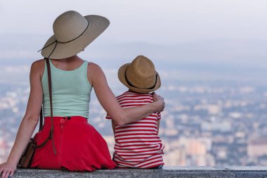Küçük oğlanı kucaklayan genç bir kadın. Anne ve oğlu şehrin panoramasına bakıyor. Arka plan