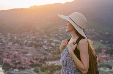 Büyük şapkalı güzel bir kadın gün batımında şehir panoramasında duruyor..