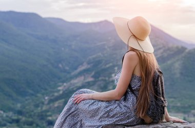 Hasır şapkalı kız tepede oturur ve dağlara bakar. Sakin dinlenme ve rahatlama kavramı