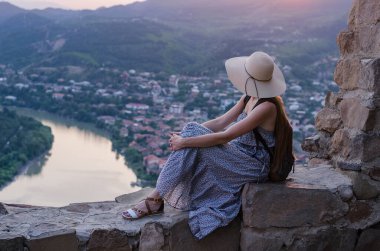 Şapkalı kız nehir ve şehrin arka planında tepede oturuyor. Sakin ol ve rahatla