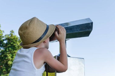 Mavi gökyüzü arka planında teleskopla turist arayan hasır şapkalı çocuk.