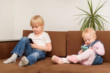 Erkek ve kız kardeş kanepede oturup telefona bakıyorlar. Sarı saçlı kardeşler. Jenerasyon Alfa kavramı.