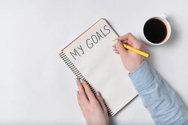 Female hands write words MY GOALS on notebook. White background. Morning coffee and plans.