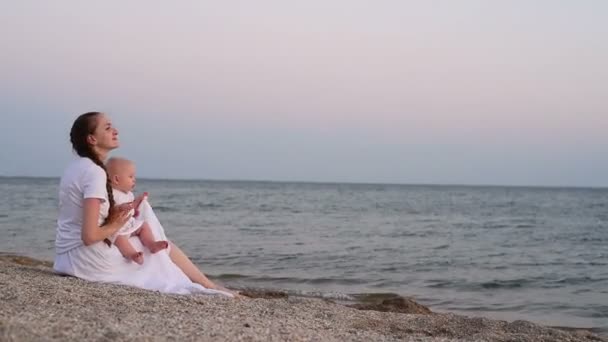 身穿白色长裙的母亲与坐在海滩上的女婴交谈 年轻的母亲带着小女儿在海上 — 图库视频影像