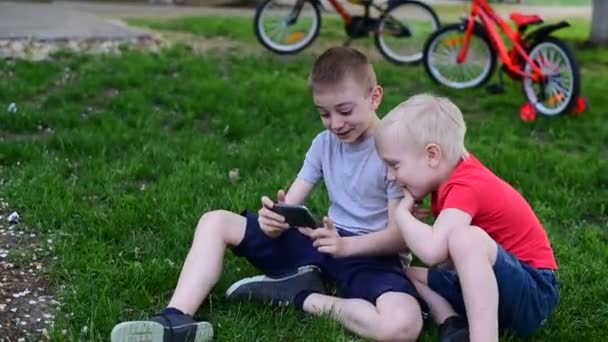 2人の男の子の友人は スマートフォンを見て 芝生の上に座って楽しい笑い ブロンドと黒髪の男の子が屋外で楽しんでいます — ストック動画
