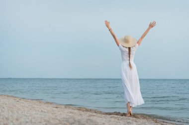 Hasır şapkalı ve beyaz elbiseli genç bir kadın kumsalda yürüyor ve ellerini havaya kaldırıyor..