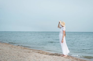 Uzun beyaz elbiseli, narin bir kadın denizin kenarında duruyor.