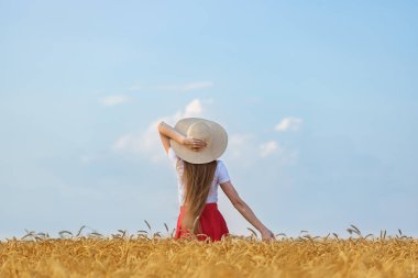 Şapkalı genç kadın mavi gökyüzü arka planındaki buğday tarlasında duruyor. Hafta sonu açık hava.
