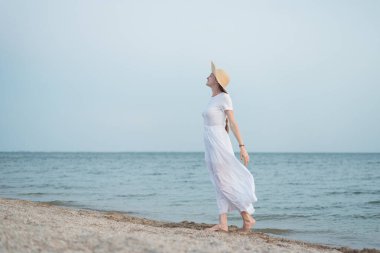 Plajda beyaz elbiseli ve şapkalı güzel bir kadın. Deniz kenarındaki aristokrat kız.