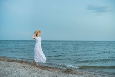 Uzun beyaz elbiseli aristokrat kadın sahilde yürüyor. Deniz tatili.