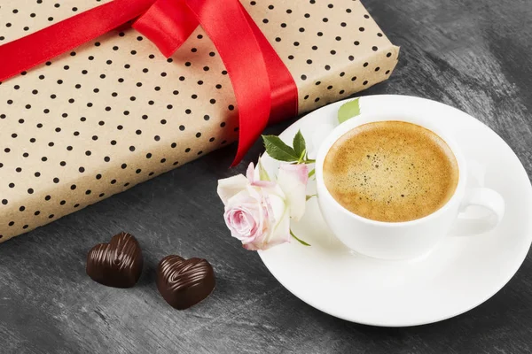 Espresso-Kaffee in einer weißen Tasse, eine rosa Rose, ein Geschenk mit rotem Band und Pralinen auf dunklem Hintergrund. — Stockfoto