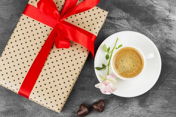 Espresso-Kaffee in einer weißen Tasse, eine rosa Rose, ein Geschenk mit rotem Band und Pralinen auf dunklem Hintergrund. Ansicht von oben. Lebensmittel-Hintergrund. — Stockfoto
