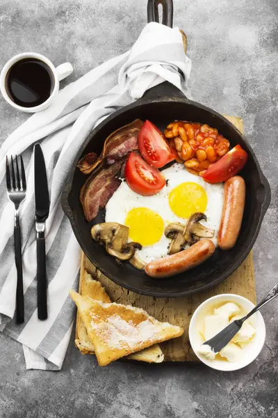 传统的英国早餐-培根, 香肠, 煎蛋, 西红柿, 豆类, 蘑菇, 烤面包用 butter-in 烹饪锅和咖啡的灰色背景。顶部视图。食品背景 — 图库照片