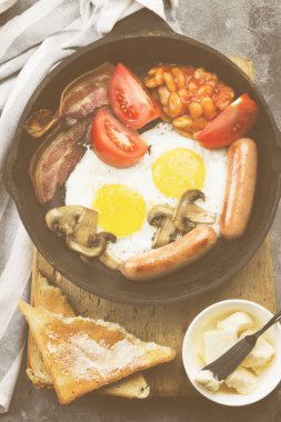 Geleneksel İngiliz kahvaltısı - pastırma, sosis, sahanda yumurta, domates, fasulye, mantar, tereyağı tava ve gri arka plan üzerinde kahve pişirme ile-tost. Üstten Görünüm. Gıda arka plan. Tonlama