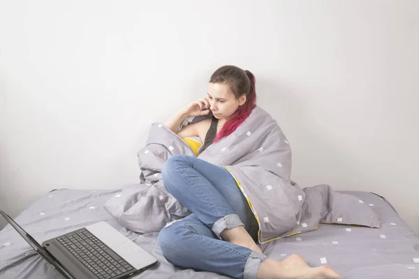 Concept - work from home, self-isolation, quarantine. Young girl sits in bed covered with blanket, looks into laptop