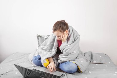 Konsept: Evden çalışma, kendini izole etme, karantina. Genç kız battaniyeyle kaplı olarak yatakta oturuyor, dizüstü bilgisayarına bakıyor.