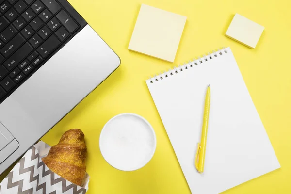 Working morning. Breakfast at work - cappuccino, croissant, notebook on yellow background. Top view