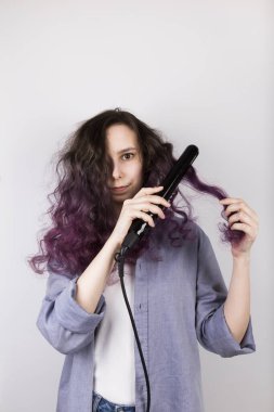 Genç kız düz demir kıvırcık saçları düzleştiriyor. Mor renkli saçlar. Gri arkaplan