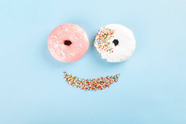 Beyaz ve pembe kremalı çörek ve mavi arka planda gülücük şeklinde çok renkli tatlı pasta. Yemekten komik surat. Üst görünüm