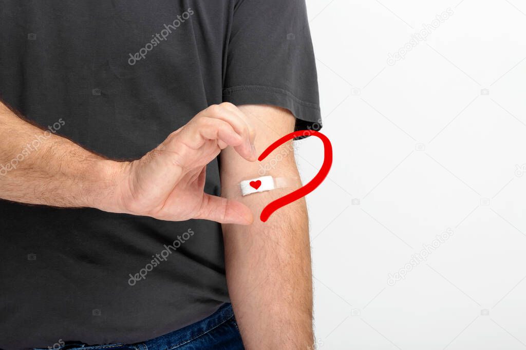 Blood donorship. Man in gray T-shirt hand taped with patch with red heart after giving blood on gray background. Shows half heart with his hand other half of the heart drawn red. Copy space