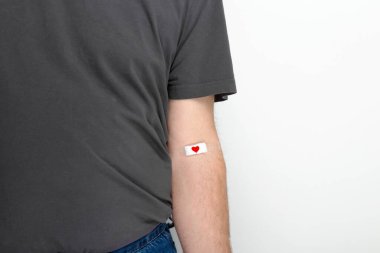 Kan bağışı. Gri arka planda kan veren gri tişörtlü adam kırmızı kalpli yamayla bantlanmış. Boşluğu kopyala