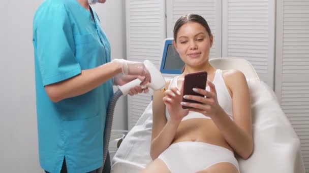 Young woman in laser epilation procedure, use smartphone. — Stock Video