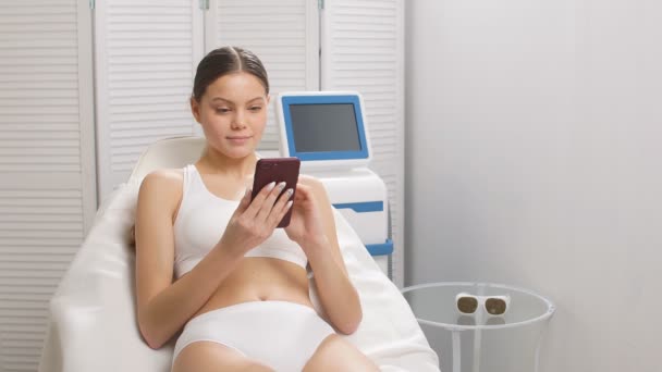 Mujer joven en el procedimiento de depilación láser, utilice el teléfono inteligente. — Vídeo de stock