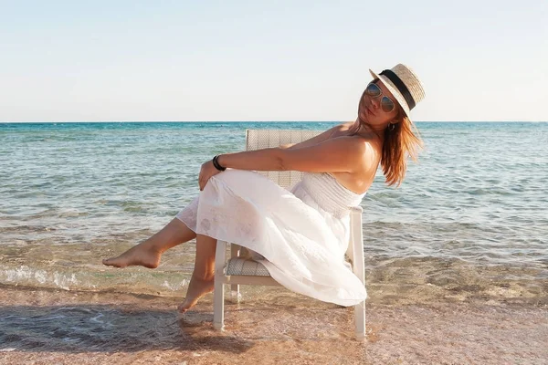Νεαρή γυναίκα με λευκό φόρεμα και καπέλο που κάθεται σε μια καρέκλα στη θάλασσα — Φωτογραφία Αρχείου