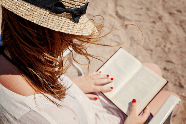 Young adult woman with a hat on the beach reading a book