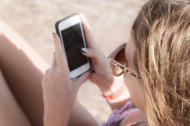 Genç bir yanık tenli kadın güneş gözlüğü ile bir akıllı telefon içine görünüyor 