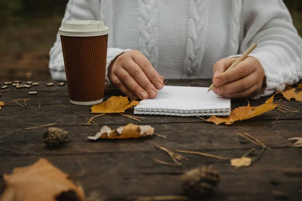 Human hands with a cup of coffee and scarf at wooden table with notebook and pen and autumn leaves. Human hands with a cup of coffee and scarf at wooden table