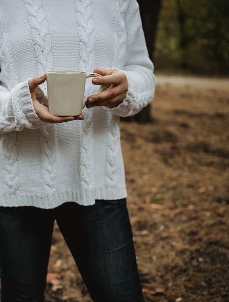 Jeune femme dans un pull blanc tient une tasse de thé avec du café sur un fond de feuilles d'automne jaunes. Ambiance automnale chaude, mise au point douce. Espace de copie — Photo